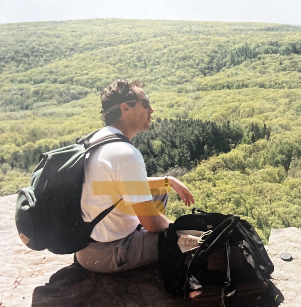 Jeffrey Berger sitting on a rocky overlook with a backpack and duffel bag, facing a wide green valley.