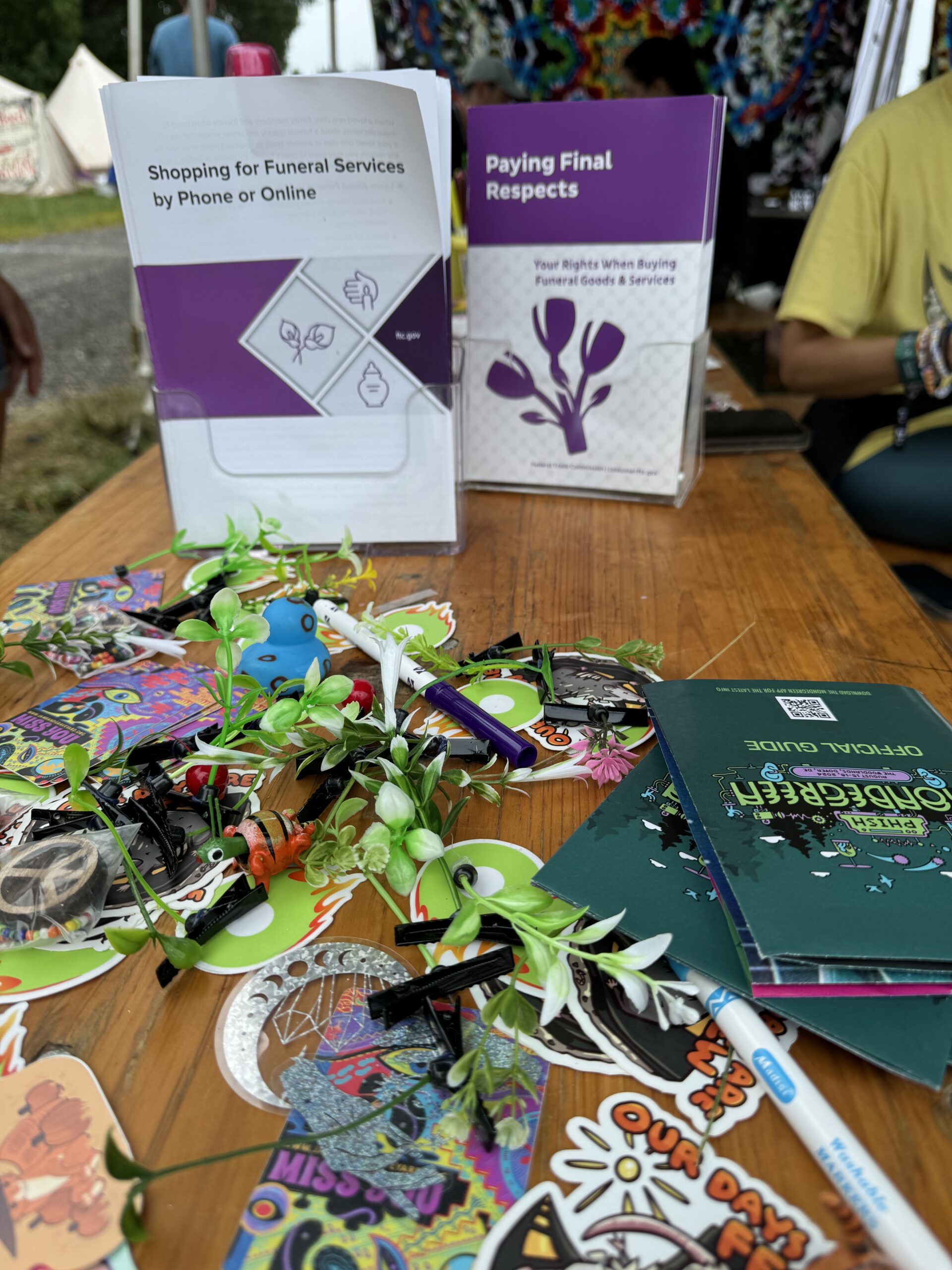 Federal Trade Commission funeral consumer rights brochures—"Shopping for Funeral Services by Phone or Online" and "Paying Final Respects"—displayed on a Jam Band Graveyard table alongside festival stickers, flowers, art supplies, and a Phish festival guide.